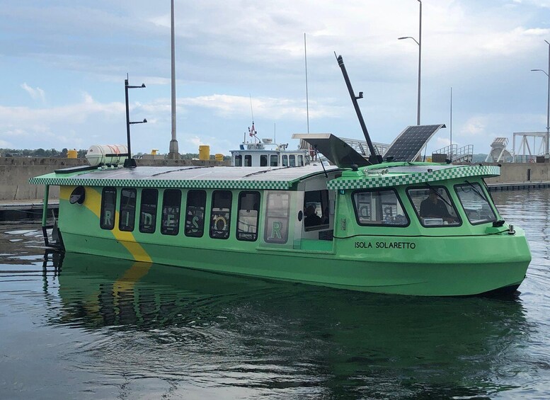 A green boat with solar panels on the roof, labeled 'ISOLA SOLARETTO,' docked at a pier with other boats and structures in the background. 2