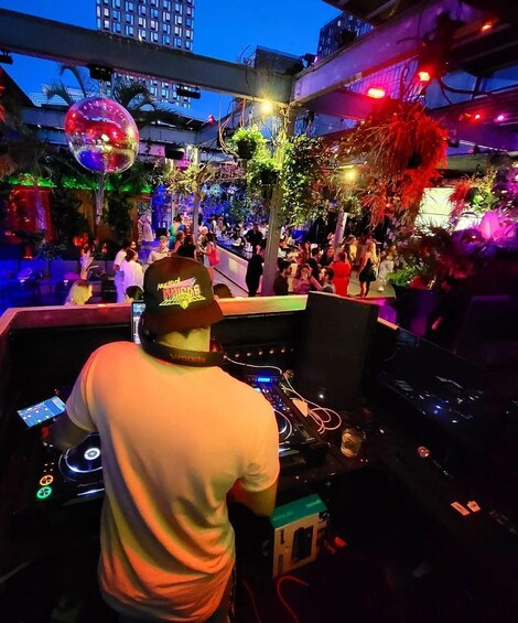 A DJ booth with electronic equipment, surrounded by a crowd at a lively outdoor event with decorative lights and palm trees. 1