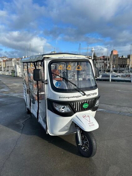 A white three-wheeled vehicle parked on a marina with a cityscape in the background. 1