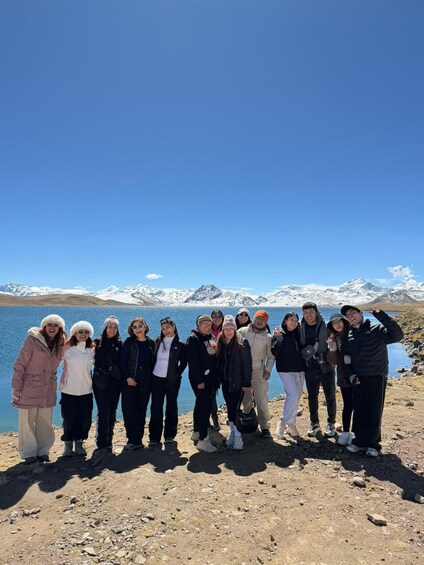A group of people standing on a rocky shore with a lake and snow-capped mountains in the background. 5