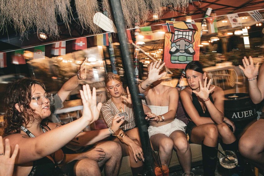 A group of people sitting on a couch with their hands raised, in a dimly lit room with flags and a poster on the wall. 1