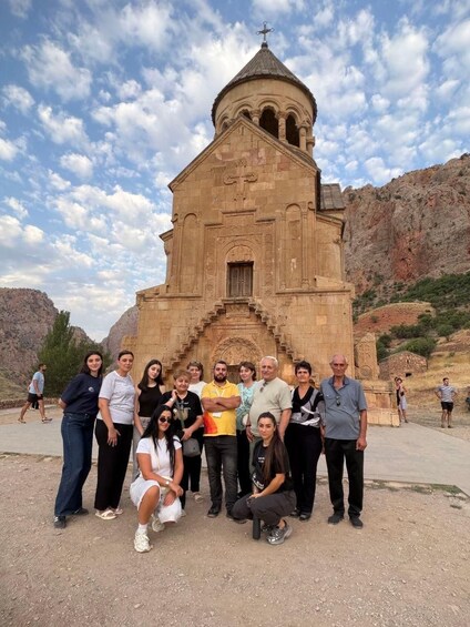 A group of people posing in front of a stone church with a cross on top, set against a mountainous backdrop. 2