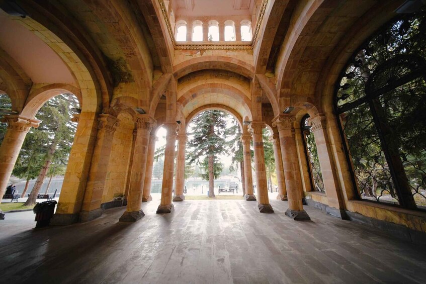 A covered colonnade with arches and columns, leading to a view of trees and a distant building. 4