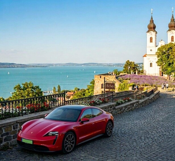 A red sports car parked on a cobblestone street, overlooking a body of water with sailboats, a church with two domes, and a building labeled 'Echo'. 1