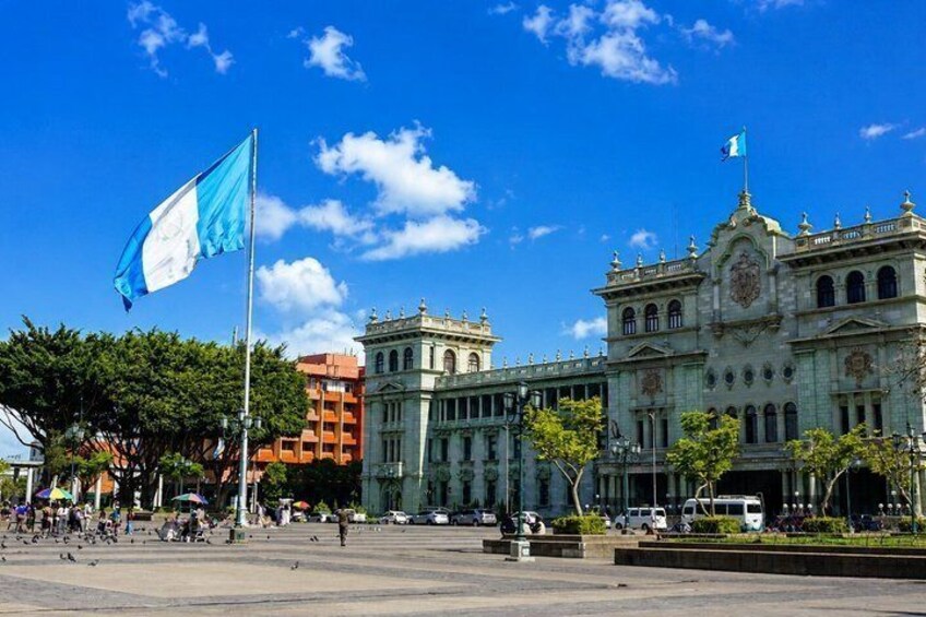 A historic building with a flag, a clear blue sky, and a plaza with trees and people. 3