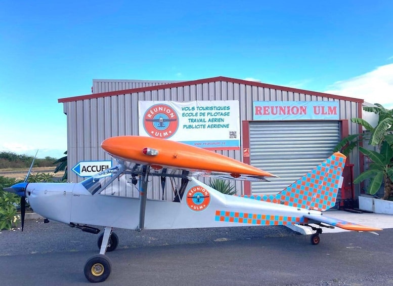 A small orange and white airplane parked in front of a building with a sign that reads 'REUNION ULM'. 2