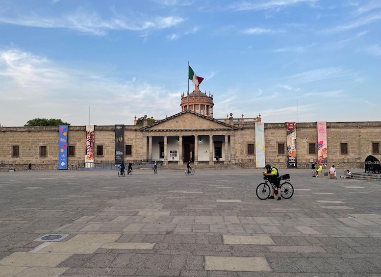 A historic building with a flag, banners, and a paved plaza with cyclists. 5
