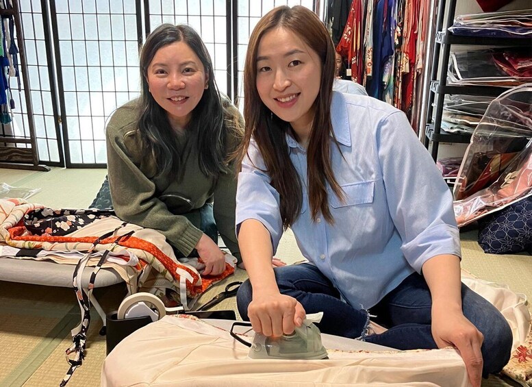 Two people ironing clothes on a tatami mat in a room with traditional Japanese sliding doors and clothing racks. 2