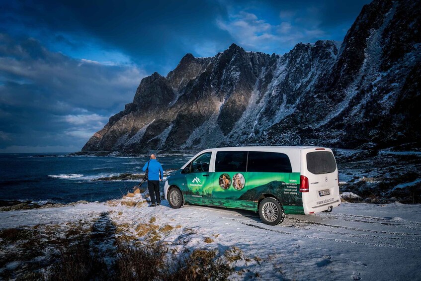 A green and white van parked on a snow-covered path with a person standing beside it, overlooking a rugged coastline and mountains under a partly cloudy sky. 3
