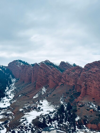 A mountainous landscape with red rock formations, snow patches, and a building in the foreground. 3