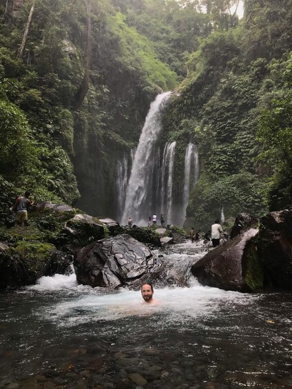 A waterfall surrounded by lush greenery, with people standing on rocks and a person in the water. 1