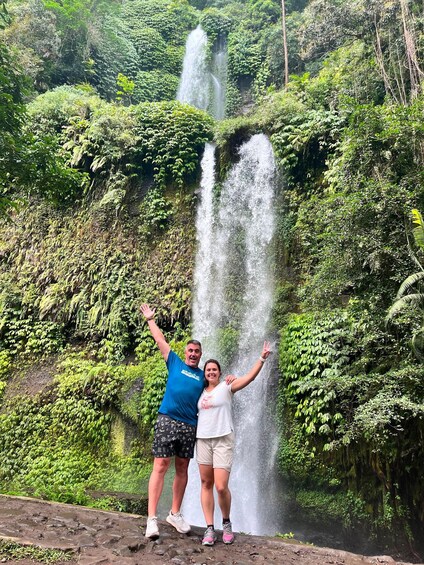 A waterfall with lush greenery and two people standing in front of it. 4