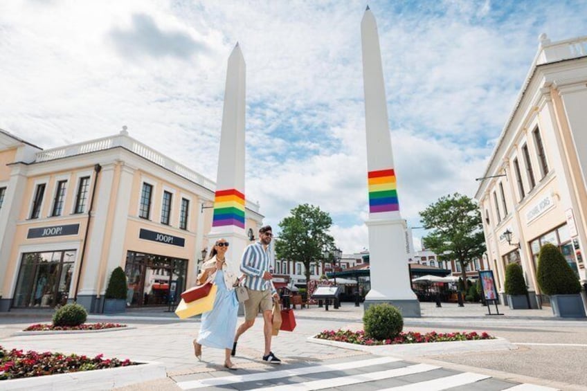 Two obelisks with rainbow stripes, a crosswalk, and storefronts with 'JOOP!' signage. 3