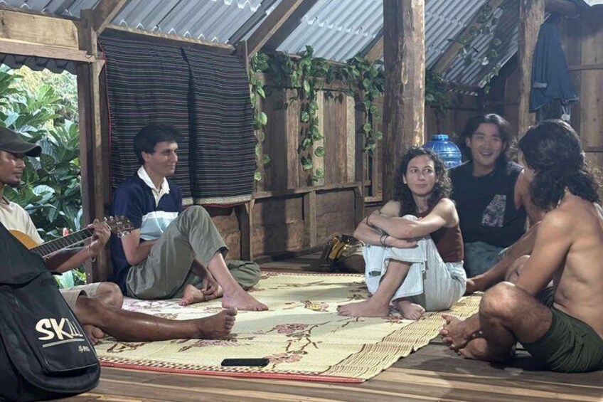 A group of people sitting on a mat in a rustic outdoor setting with wooden structures and greenery in the background. 2