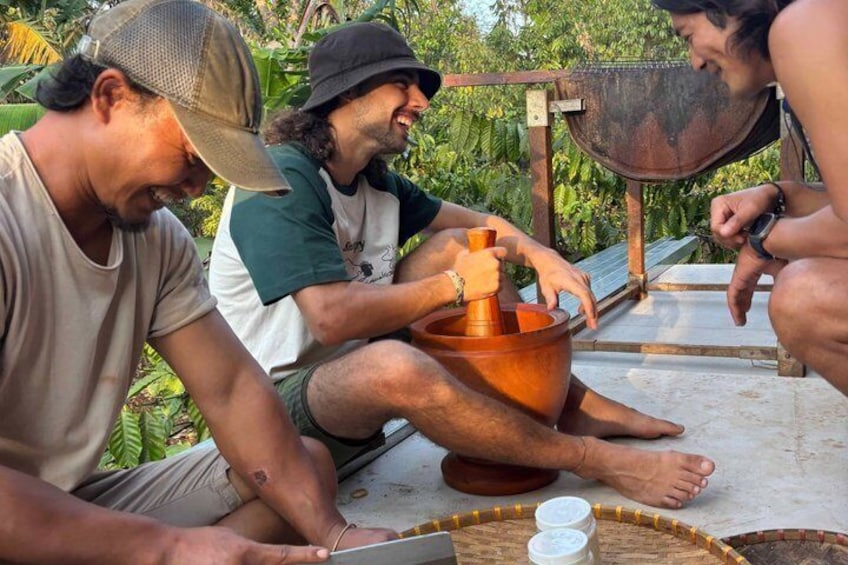 Three people sitting outdoors, one using a mortar and pestle, with a wooden table and a metal bowl in the foreground. 4