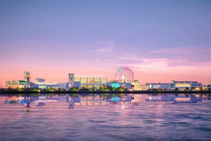 A waterfront with a Ferris wheel, modern buildings, and a calm body of water reflecting the structures. 3