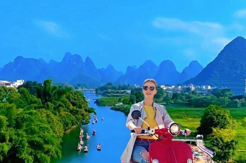 A person on a red motorcycle, a river, and green hills with mountains in the background. 1