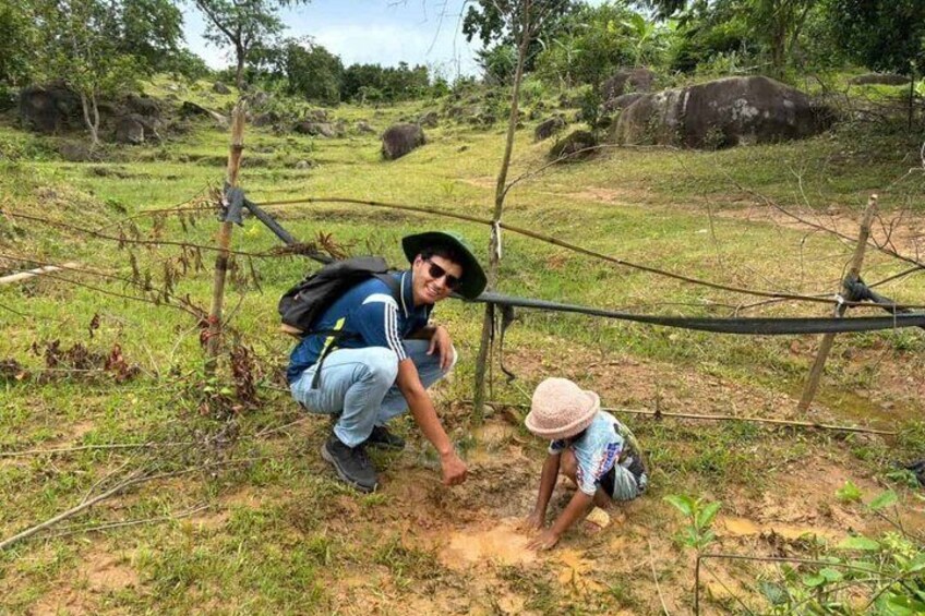 A person in a blue shirt and black hat crouching next to a child in a patterned shirt and pink hat, both near a makeshift rope bridge in a grassy area with trees and rocks in the background. 5