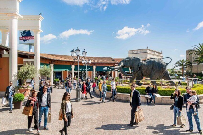 A public square with a large elephant statue, people walking, and buildings with signs. 4