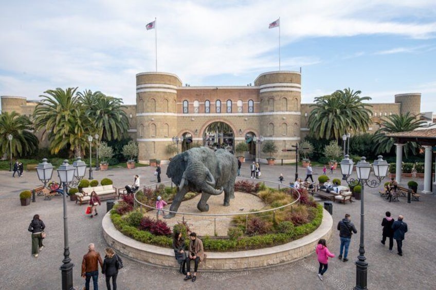 A large elephant statue in a plaza with people walking around, palm trees, and a building with a flag. 2