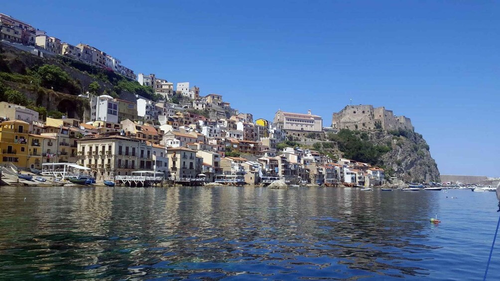 A coastal town with buildings on a hillside, a fort on a cliff, and boats in the water. 2