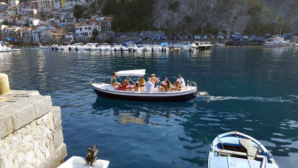 A boat with passengers on a calm body of water, surrounded by a coastal town with buildings and boats docked along the shore. 1