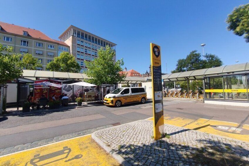 A bus stop with a yellow sign, a parked van, and a building in the background. 5