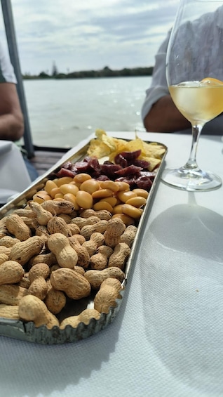A tray of snacks including peanuts, almonds, and dried fruits, and a glass of white wine on a table by a body of water. 4