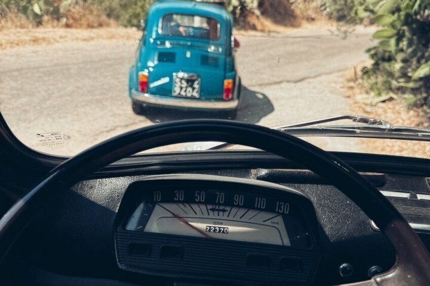 A car dashboard with a speedometer, a steering wheel, and a view of a road with a blue car ahead. 2