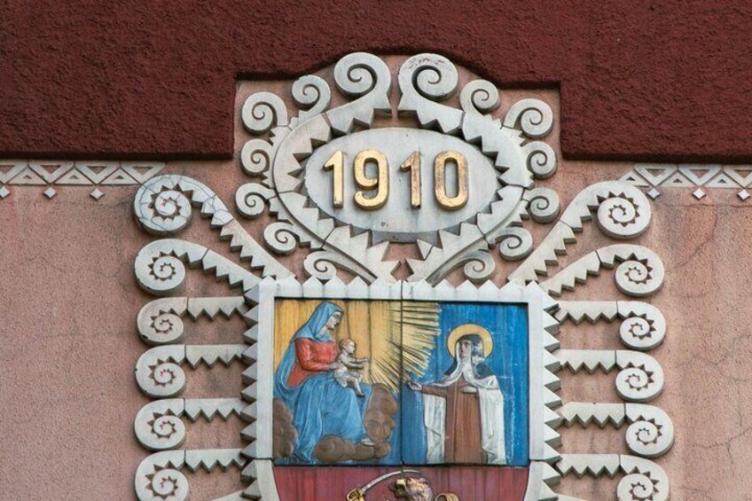 A building facade with a decorative metal sign displaying the year 1910, surrounded by intricate patterns and a mural depicting a religious scene. 3
