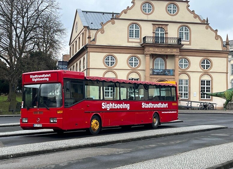 A red sightseeing bus with 'Stadtverkehr' and 'Sightsseeing' text on the side, parked in front of a historic building with arched windows and a balcony. 5