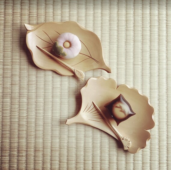 Two decorative leaf-shaped objects with a small, round item on one and a square item on the other, placed on a textured surface. 2