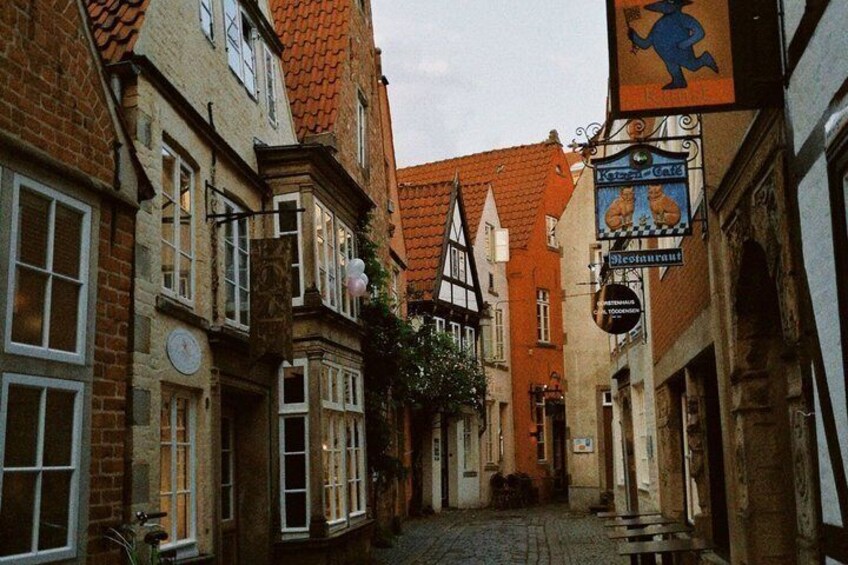 A narrow cobblestone street lined with traditional half-timbered buildings, a sign for a local bakery, and a clock on one of the buildings. 3