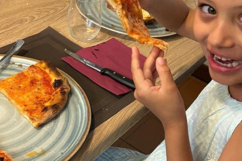 A child holding a slice of pizza, a knife, and a glass of water on a table. 3