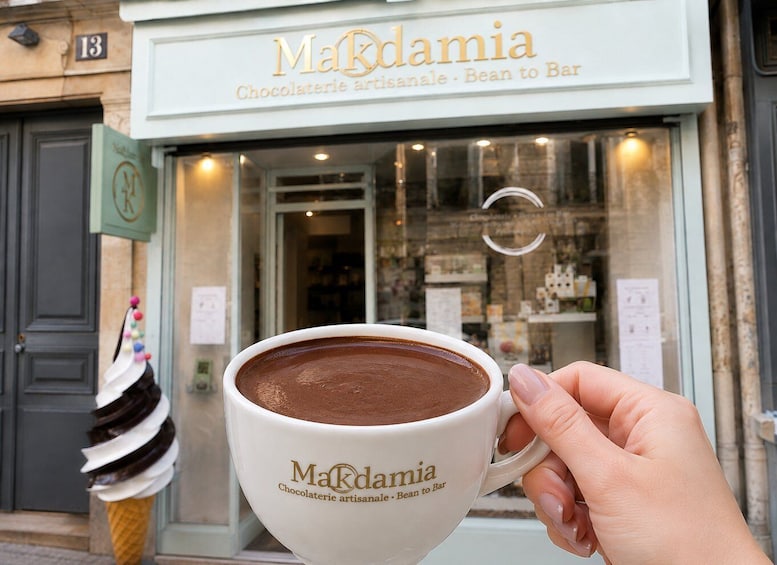 A hand holding a cup of hot chocolate with the 'Makdamia' logo, in front of a shop with a sign reading 'Makdamia Chocolaterie artisanale - Bean to Bar'. 4