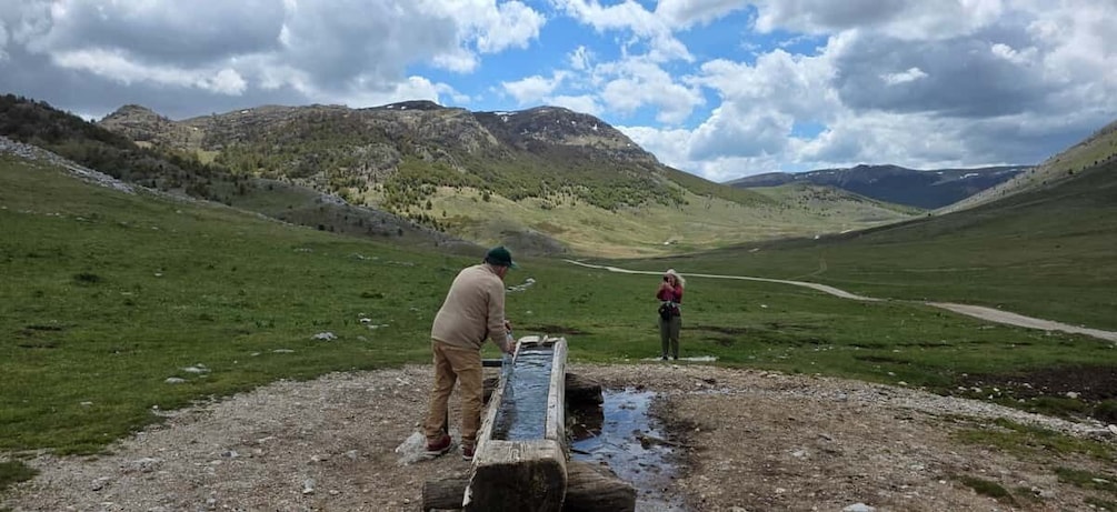 Two people standing by a wooden trough in a grassy valley with mountains in the background. 3
