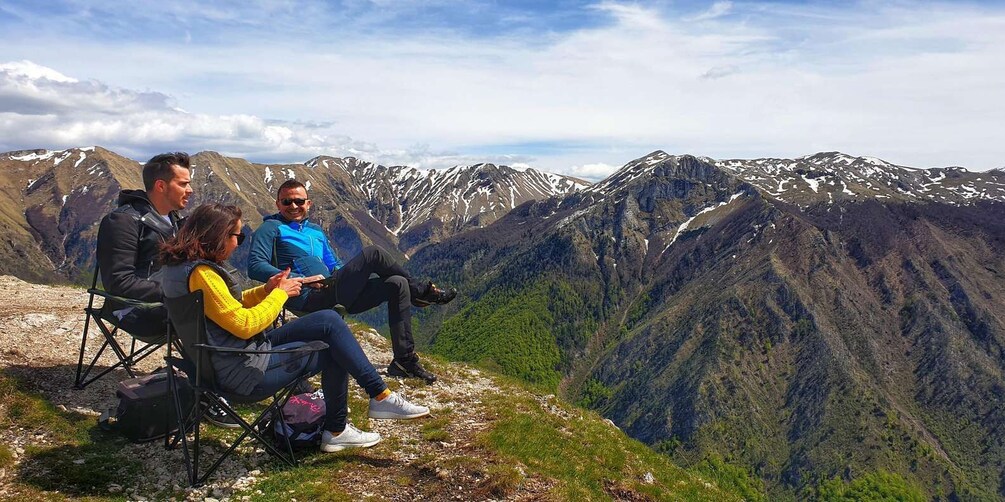 Three people sitting on a mountain ledge with a view of a valley and snow-capped mountains. 1