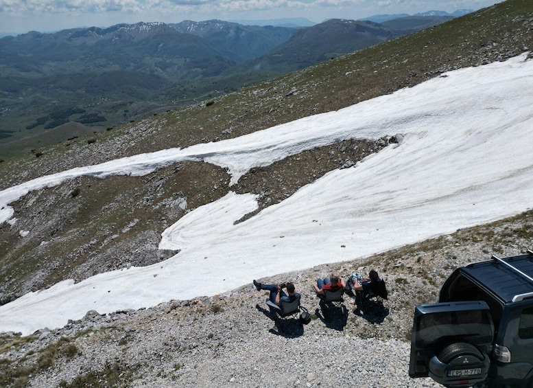 A snowy mountain landscape with a group of people sitting on chairs, a parked vehicle, and a clear sky. 2