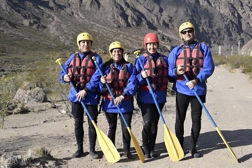 Four people in blue jackets and helmets holding paddles, standing on a dirt path with rocky terrain and mountains in the background. 4