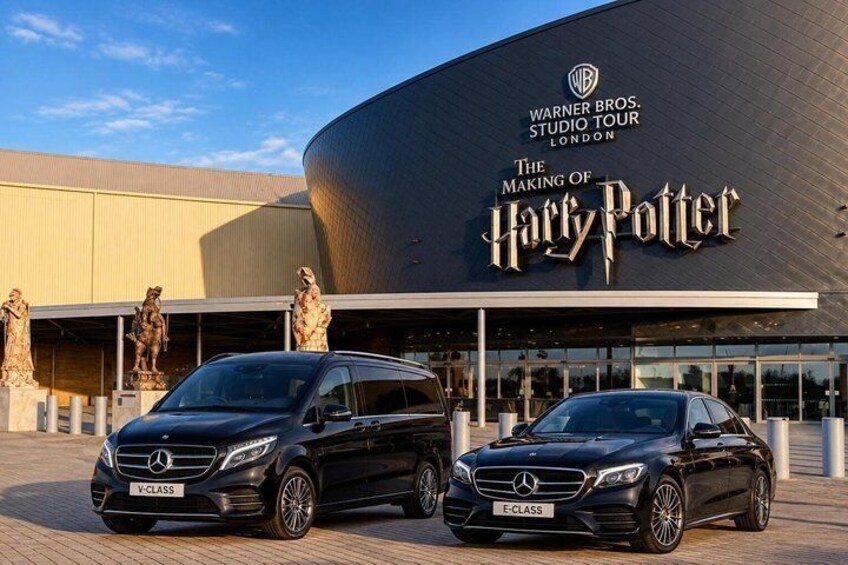 A building with a curved facade and the Warner Bros. Studio Tour London sign. Two black Mercedes cars are parked in front. 3