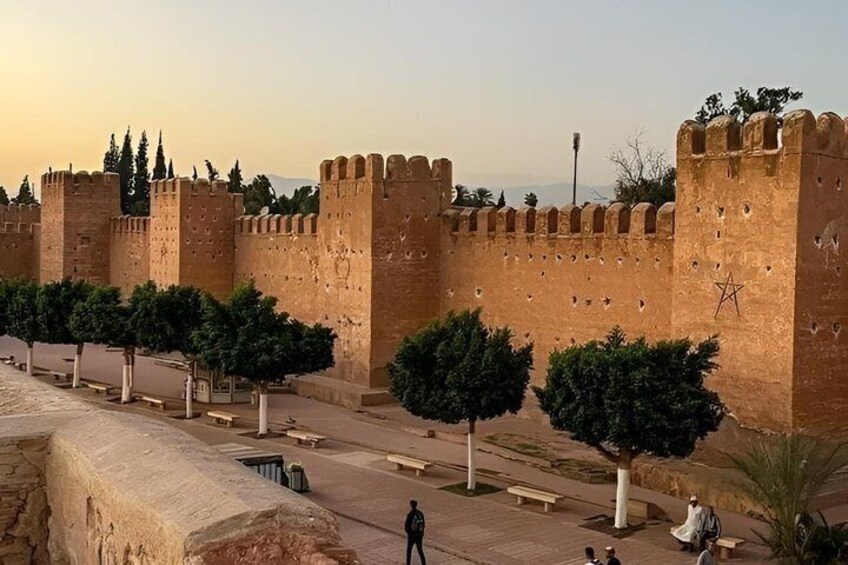 A historic fort with large stone walls and towers, a paved walkway, and trees. 2