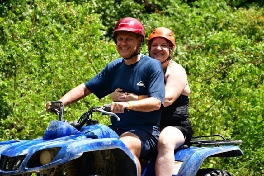 Two people on an ATV in a green, leafy area. 1