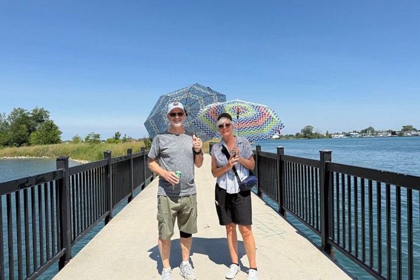 Two people on a bridge with a railing, holding umbrellas, and a body of water in the background. 2