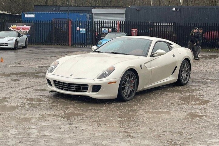 A white sports car parked on a wet surface with a fence and another car in the background. 2
