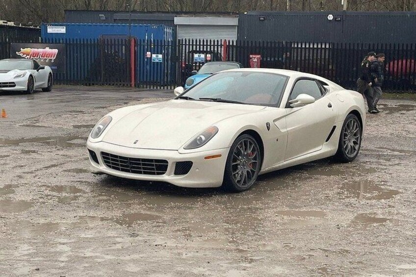 A white sports car parked on a wet surface with a fence and another car in the background. 2