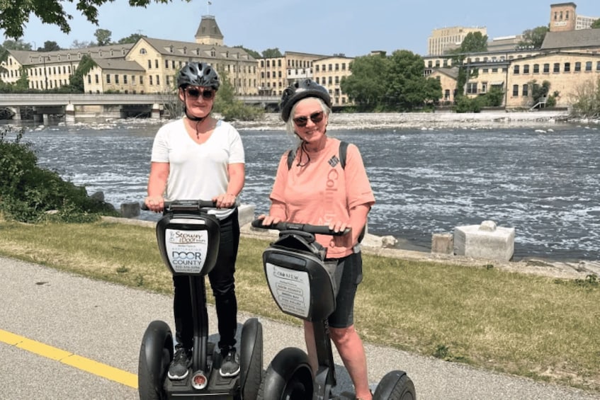 Two people on Segway devices near a river with buildings in the background. 1