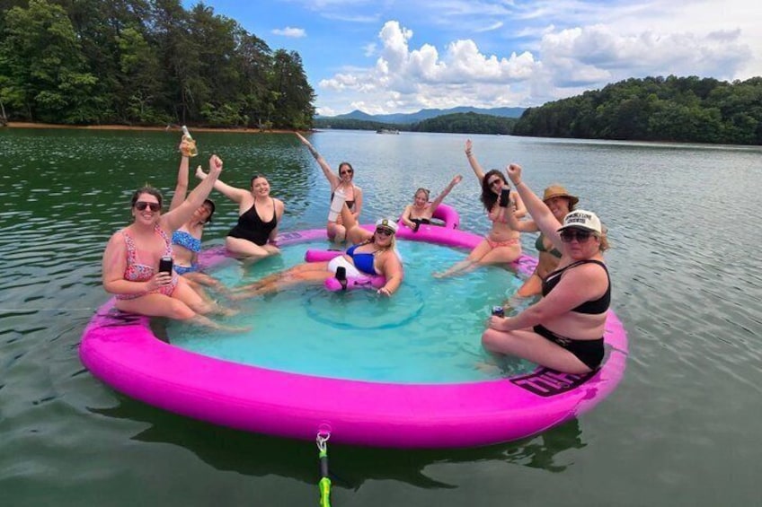 A group of people on a pink inflatable raft in a lake surrounded by trees and hills. 1