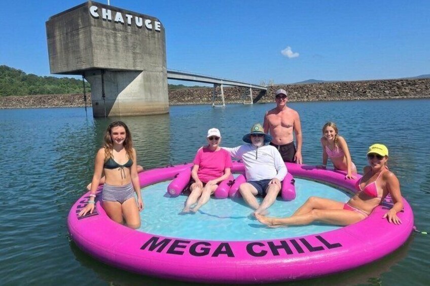A group of people on a pink inflatable pool with 'MEGA CHILL' written on it, floating on water with a concrete structure labeled 'CHATUGE' in the background. 2