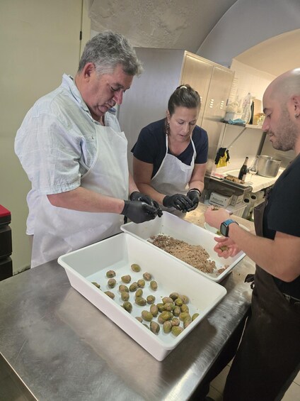 Three people in a kitchen preparing food, with trays of olives and a bottle of wine on the counter. 4