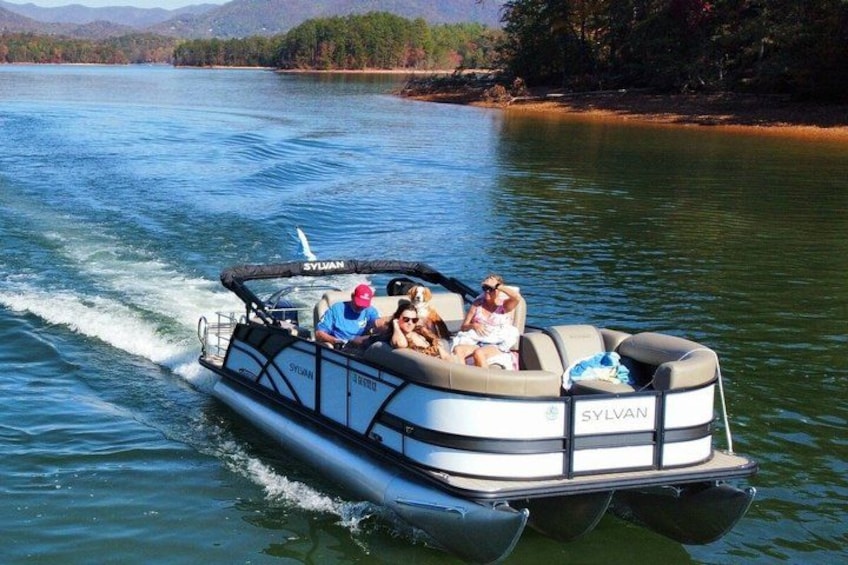 A boat with the brand 'SYLVAN' on the side, moving on a body of water with a forested shoreline in the background. 2
